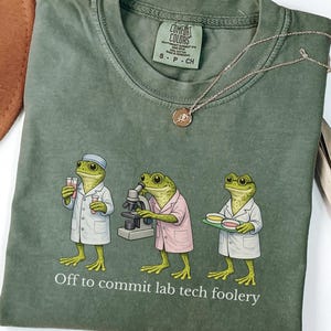 May include: Moss green t-shirt featuring a graphic of three frog scientists in lab coats, with the text "Off to commit lab tech foolery." The shirt has a crew neck and short sleeves. A gold necklace with a pendant is draped across the shirt.