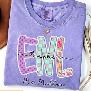 May include: A violet Comfort Colors t-shirt with a graphic that says "EML teacher" and "Ms. Miller". The letters are decorated with polka dots, a floral pattern, and a checkered pattern. A gold necklace is draped over the shirt.