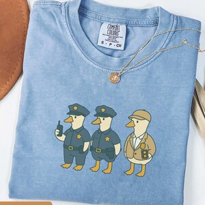 May include: Blue jean colored t-shirt featuring a cartoon illustration of three ducks dressed as police officers. The ducks are wearing blue uniforms and hats. The shirt has a Comfort Colors tag and a gold necklace. The text "BLUE JEAN" is at the bottom.