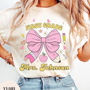 Puede incluir: Camiseta color crema con un gran lazo rosa y un gráfico de lápiz. El texto "FIRST GRADE Mrs. Johnson" está en amarillo. Gráficos adicionales incluyen corazones, estrellas, flores y una manzana. La camiseta es de estilo casual.