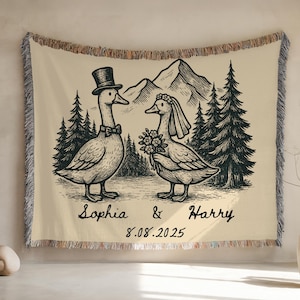 May include: A cream-colored woven throw blanket featuring a black and white illustration of two geese dressed for a wedding. The goose on the left wears a top hat and bow tie, while the goose on the right wears a veil and holds a bouquet. The background includes mountains and pine trees. The names 'Sophia & Harry' and the date '8.08.2025' are written below the geese.