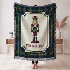 May include: A woven throw blanket featuring a dark green, blue, and red plaid pattern with a central cream-colored panel. The panel displays a nutcracker illustration and the text THE MILLERS in dark red letters, finished with fringed edges.