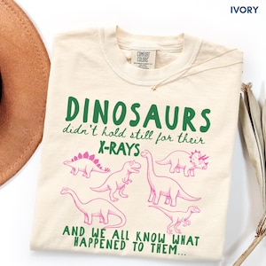 May include: An ivory t-shirt featuring green text that reads "DINOSAURS didn't hold still for their X-RAYS AND WE ALL KNOW WHAT HAPPENED TO THEM..." and pink line-art illustrations of five different dinosaurs arranged across the front.