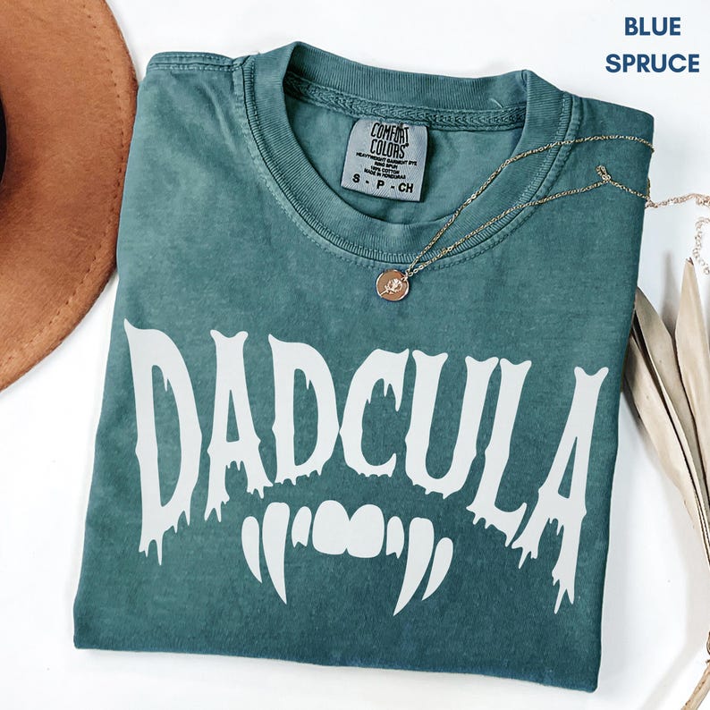 May include: A blue spruce colored t-shirt with a crew neckline and a Comfort Colors brand tag. The shirt features the white text DADCULA in a dripping font style, accompanied by a graphic of white vampire fangs centered below the text.