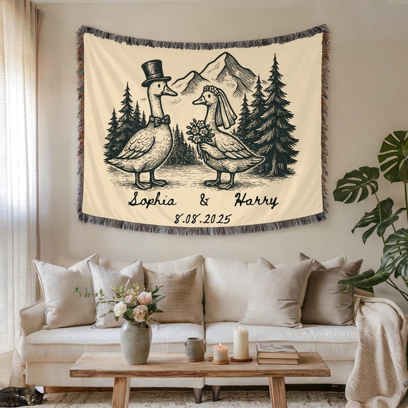 May include: Cream-colored woven throw blanket featuring a black and white illustration of two geese dressed for a wedding. The blanket has a fringed edge, with the names "Sophia & Harry" and the date "8.08.2025" printed below the geese.