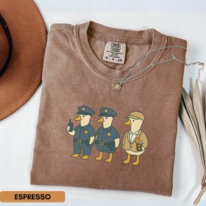 Może przedstawiać: Koszulka w kolorze espresso z zabawną ilustracją trzech postaci kaczek przebranych za policjant&oacute;w i detektywa. Koszulka ma okrągły dekolt i kr&oacute;tkie rękawy. Srebrny naszyjnik z wisiorkiem jest przerzucony przez koszulkę.