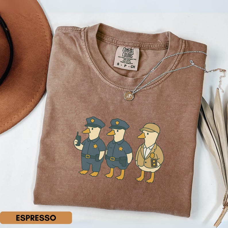 Może przedstawiać: Koszulka w kolorze espresso z zabawną ilustracją trzech postaci kaczek przebranych za policjant&oacute;w i detektywa. Koszulka ma okrągły dekolt i kr&oacute;tkie rękawy. Srebrny naszyjnik z wisiorkiem jest przerzucony przez koszulkę.