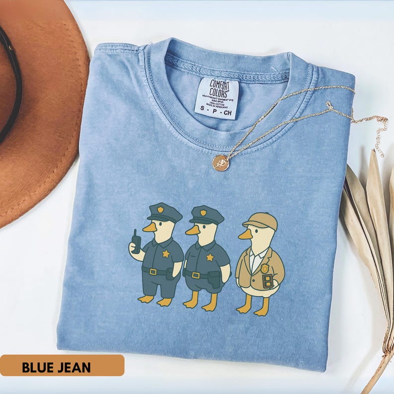 May include: Blue jean colored t-shirt featuring a cartoon illustration of three ducks dressed as police officers. The ducks are wearing blue uniforms and hats. The shirt has a Comfort Colors tag and a gold necklace. The text "BLUE JEAN" is at the bottom.