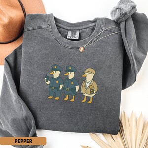 Może przedstawiać: Ciemnoszara bluza z zabawną ilustracją trzech postaci kaczek w mundurach policjant&oacute;w. Jedna kaczka trzyma kr&oacute;tkofal&oacute;wkę. Słowo "PEPPER" jest wydrukowane na małej, prostokątnej etykiecie. Okrągły dekolt i długie rękawy.