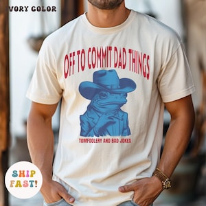 May include: Ivory-colored t-shirt with the text "OFF TO COMMIT DAD THINGS" in red. The shirt features a blue frog wearing a cowboy hat and suit, holding a cigar. The text "TOMFOOLERY AND BAD JOKES" is below the frog.