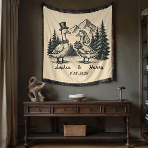 May include: Cream-colored tapestry with a black and white illustration of two geese dressed for a wedding, with a mountain and forest backdrop. The names "Sophia & Harry" and the date "8.08.2025" are written below the geese.
