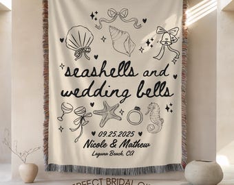 May include: Cream-colored woven throw blanket with black line art of seashells, wedding bells, and other wedding-themed illustrations. The text reads "seashells and wedding bells" with a date and names. The bottom text says "PERFECT BRIDAL GIFT".