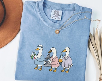 May include: Blue jean colored t-shirt with a graphic of three cartoon ducks in lab coats, holding beakers and a microscope. The shirt has a crew neck and short sleeves. A gold necklace with a pendant is draped on the shirt.