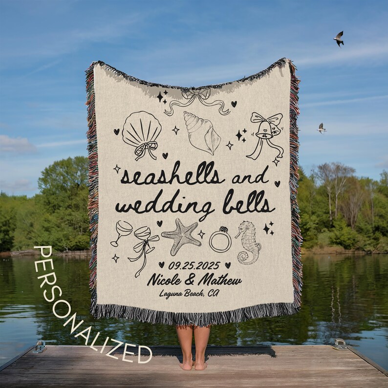 May include: A cream-colored personalized woven blanket with black illustrations of seashells, wedding bells, and other wedding-themed graphics. The blanket includes the text "seashells and wedding bells", a date, and names.