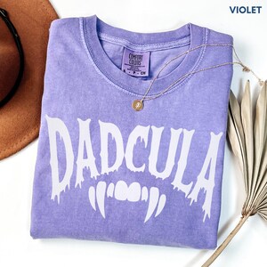 May include: A violet short-sleeved t-shirt with a crew neckline and a white graphic print that reads DADCULA with vampire fangs underneath. The shirt features a Comfort Colors label at the inner neck and is displayed with a gold-toned necklace.