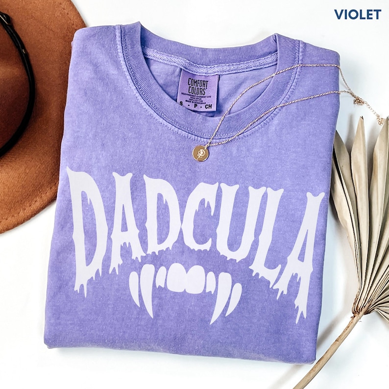 May include: A violet short-sleeved t-shirt with a crew neckline and a white graphic print that reads DADCULA with vampire fangs underneath. The shirt features a Comfort Colors label at the inner neck and is displayed with a gold-toned necklace.