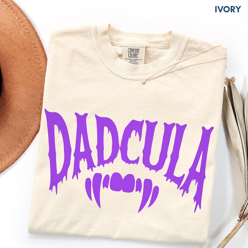 May include: An ivory short-sleeve t-shirt featuring the purple text DADCULA in a dripping, stylized font. Below the text are two purple vampire fangs. The shirt has a crew neckline and a Comfort Colors brand label printed on the inside collar.