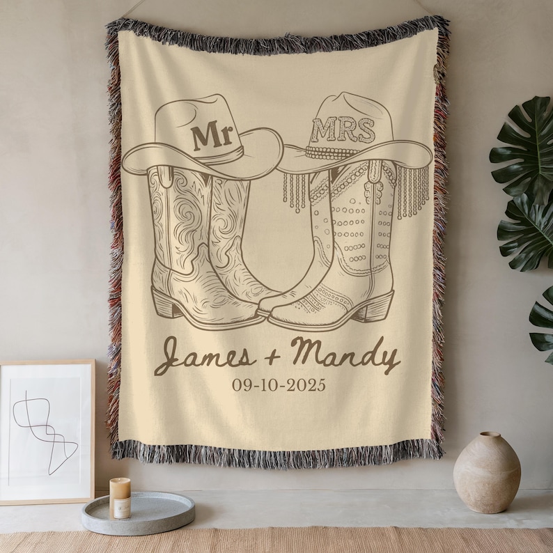 Pu&ograve; includere: Una coperta beige con un disegno a tratto di stivali e cappelli da cowboy. I cappelli sono etichettati "Mr" e "Mrs". La coperta include anche i nomi "James + Mandy" e la data "09-10-2025".