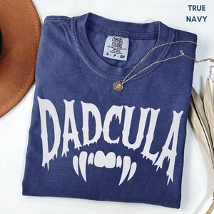 May include: A true navy blue short-sleeve t-shirt with a Comfort Colors label at the neck. The front features the white text DADCULA in a dripping font style, positioned above a graphic of white vampire fangs. A gold-toned necklace is present.