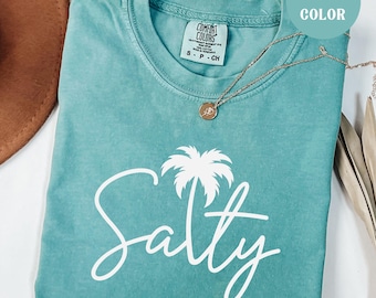 May include: Seafoam green Comfort Colors t-shirt with the word "Salty" in white script and a white palm tree graphic. A gold necklace with a pendant is draped across the shirt. The shirt is labeled "Seafoam Color".