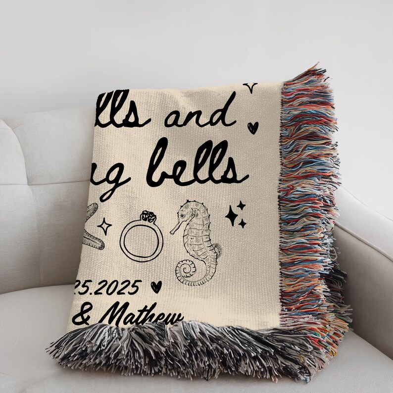 May include: Cream-colored woven throw blanket with black fringe. The blanket features the text "Bells and Ring Bells" with a ring, seahorse, and date graphic. The date "25.2025" and names are also printed on the blanket.