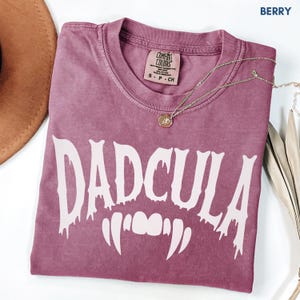 May include: A berry-colored short-sleeve t-shirt with a crew neckline. The shirt features the white text DADCULA in a dripping font style, positioned above a graphic of white vampire fangs. The interior neck label reads Comfort Colors.
