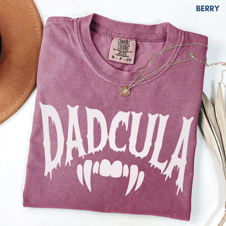 May include: A berry-colored short-sleeve t-shirt with a crew neckline. The shirt features the white text DADCULA in a dripping font style, positioned above a graphic of white vampire fangs. The interior neck label reads Comfort Colors.