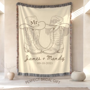 Pu&ograve; includere: Coperta tessuta color crema con un design di stivali e cappelli da cowboy. I cappelli riportano "Mr" e "Mrs". Sono visualizzati anche i nomi "James + Mandy" e la data "09-10-2025". La coperta ha un bordo sfrangiato.