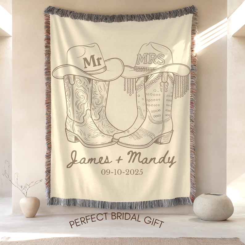 Pu&ograve; includere: Coperta tessuta color crema con un design di stivali e cappelli da cowboy. I cappelli riportano "Mr" e "Mrs". Sono visualizzati anche i nomi "James + Mandy" e la data "09-10-2025". La coperta ha un bordo sfrangiato.