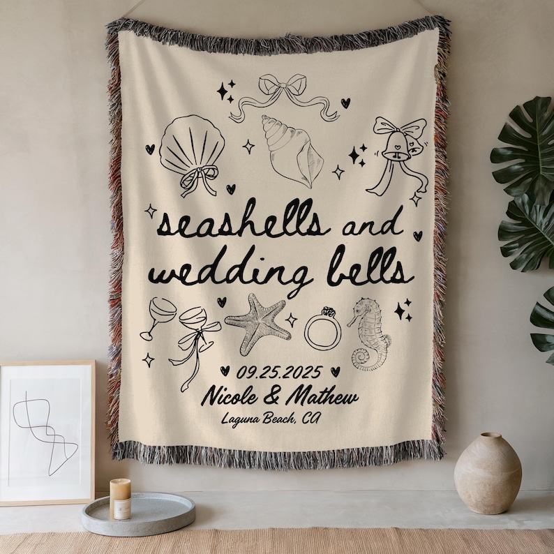May include: Cream-colored woven throw blanket with black line art of seashells, wedding bells, and other wedding-themed illustrations. The text "seashells and wedding bells" is displayed, along with a date and names.