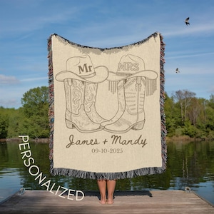 Pu&ograve; includere: Una coperta beige personalizzata con stivali e cappelli da cowboy, con le parole "Mr" e "Mrs". La coperta include anche i nomi "James + Mandy" e la data "09-10-2025".