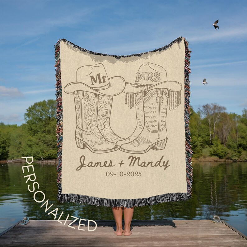 Pu&ograve; includere: Una coperta beige personalizzata con stivali e cappelli da cowboy, con le parole "Mr" e "Mrs". La coperta include anche i nomi "James + Mandy" e la data "09-10-2025".