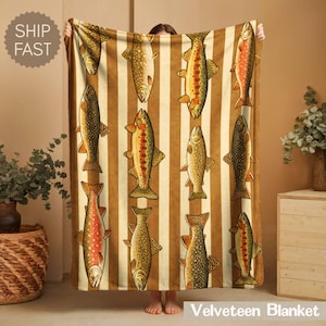 May include: A Velveteen blanket featuring a repeating pattern of illustrated fish in shades of orange, brown, and yellow against a striped background. The blanket is held up by a person. The text "Velveteen Blanket" is at the bottom.