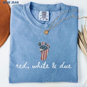 May include: A light blue Comfort Colors t-shirt with a patriotic foot graphic and the words "red, white & due". The shirt has a crew neck and short sleeves. A gold necklace with a pendant is draped over the shirt.