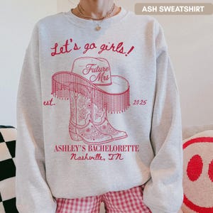 Op de afbeelding: Asgrijze sweatshirt met een rood grafisch ontwerp. Het ontwerp bevat de tekst "Let's go girls!" boven een cowboyhoed en laarzen, met "Future Mrs" en "2025" op de hoed. Daaronder staat "ASHLEY'S BACHELORETTE Nashville, TN".