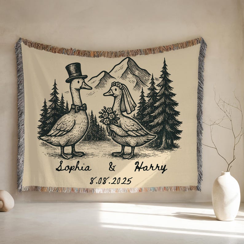 May include: A cream-colored woven throw blanket featuring a black and white illustration of two geese dressed for a wedding. The goose on the left wears a top hat and bow tie, while the goose on the right wears a veil and holds a bouquet. The background includes mountains and pine trees. The names 'Sophia & Harry' and the date '8.08.2025' are written below the geese.