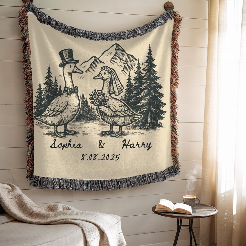 May include: Cream-colored throw blanket with a black and white illustration of two geese dressed for a wedding, with mountains and trees in the background. The blanket has a fringed edge and the names "Sophia & Harry" and the date "8.08.2025" are printed on it.