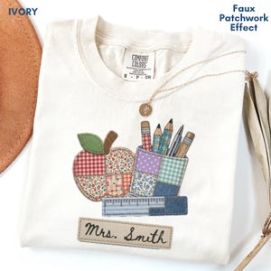 May include: An ivory t-shirt featuring a faux patchwork effect graphic of an apple, a pencil holder with various writing utensils, and a ruler. Below the graphic is a rectangular patch with the text "Mrs. Smith" printed in black script.