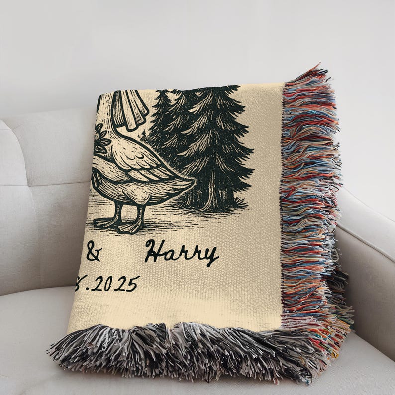 May include: Cream-colored throw blanket with a black illustration of a goose, trees, and the names "& Harry" with the date "8.2025". The blanket has a fringed edge with multiple colors.
