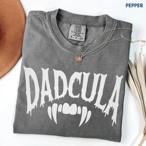 May include: A pepper-colored short-sleeve t-shirt featuring the white text DADCULA in a dripping font style with a graphic of vampire fangs underneath. The shirt has a crew neckline and a Comfort Colors brand label on the inside collar.
