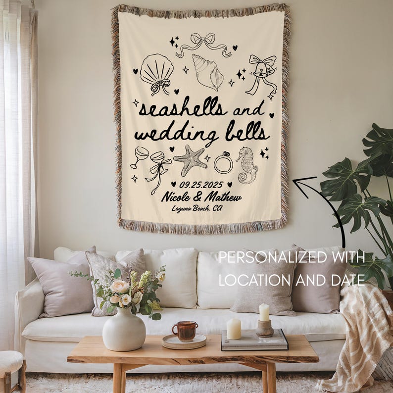 May include: Cream-colored woven throw blanket with black line art of seashells, wedding bells, and text. The text reads "seashells and wedding bells," with a date and names below. The blanket is personalized with a location and date.