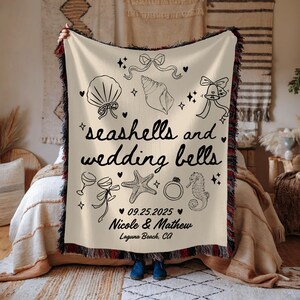 May include: Cream-colored throw blanket with black fringe and illustrations of seashells, wedding bells, and other wedding-themed graphics. Text reads "seashells and wedding bells," with a wedding date and names.