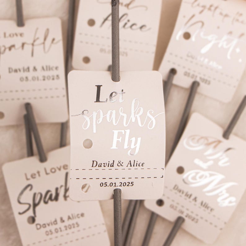 May include: Close-up of several beige sparkler tags with silver text. The tags read "Let sparks Fly," "Let Love Spark," and "Light up the Night." Each tag includes a name and date. The sparklers are gray.