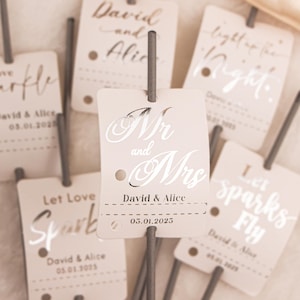 May include: Close-up of several wedding sparkler tags with text. The tags are light beige with silver text that reads "Mr and Mrs", names, and a date. Other tags say "Let Love Spark" and "Let Sparks Fly".