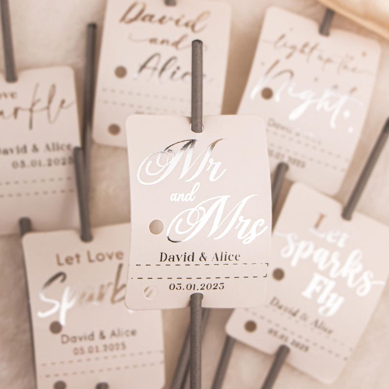May include: Close-up of several wedding sparkler tags with text. The tags are light beige with silver text that reads "Mr and Mrs", names, and a date. Other tags say "Let Love Spark" and "Let Sparks Fly".