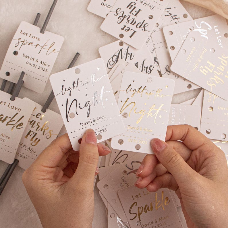 May include: A collection of white tags with gold and silver foil lettering. The tags read "Let Love Sparkle", "Let Sparks Fly", and "light up the Night". Each tag includes a date and names.