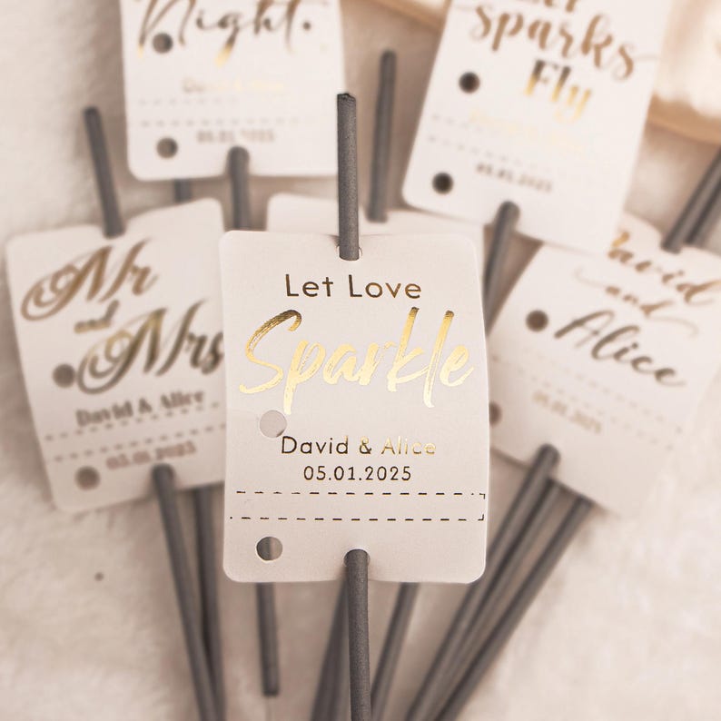 May include: A collection of wedding sparklers with white tags and gold lettering. The tags read "Let Love Sparkle", "Mr and Mrs", and "David & Alice" with a date. The sparklers have gray sticks.