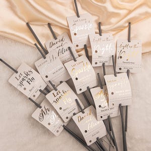 May include: A collection of wedding sparklers with attached tags. The tags are light beige with gold and silver foil text that reads "Let Love Sparkle", "Let Sparks Fly", and "light up the night". Each tag includes a date and names.