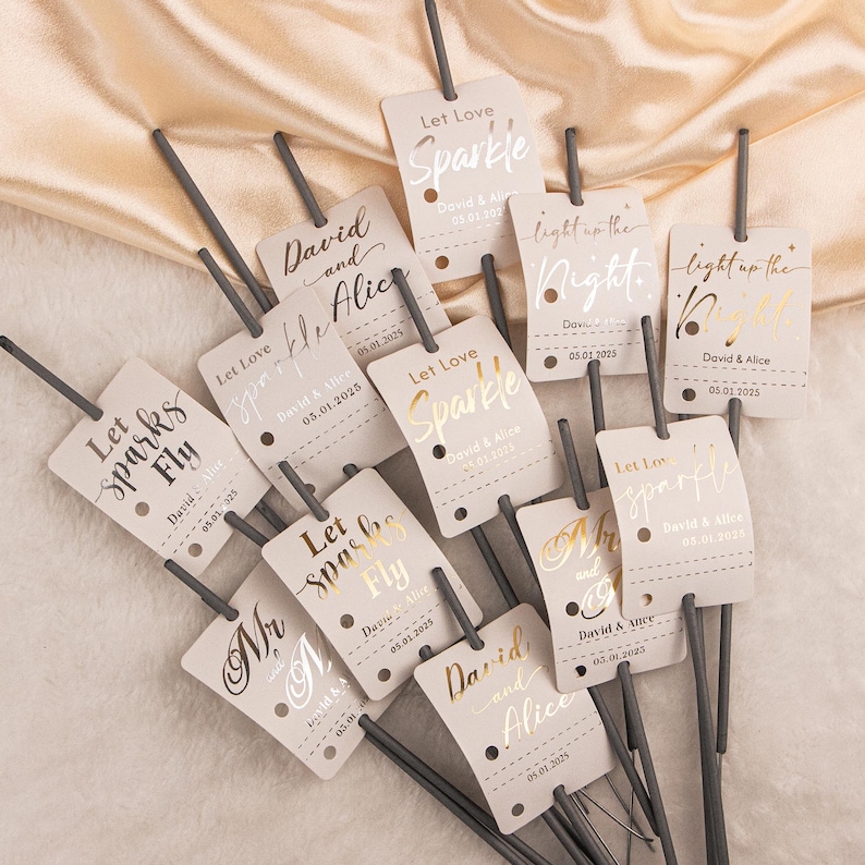 May include: A collection of wedding sparklers with attached tags. The tags are light beige with gold and silver foil text that reads "Let Love Sparkle", "Let Sparks Fly", and "light up the night". Each tag includes a date and names.