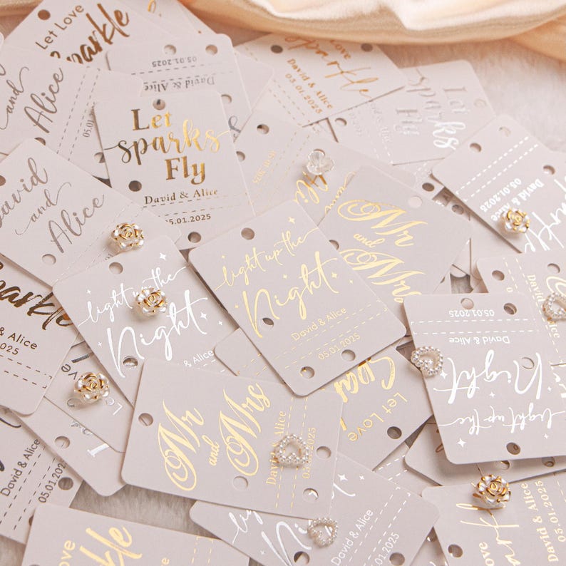 May include: A collection of light gray wedding favor tags with gold foil lettering and small decorative earrings. The tags feature phrases like "Let Sparks Fly" and "Light up the Night", along with names and dates.